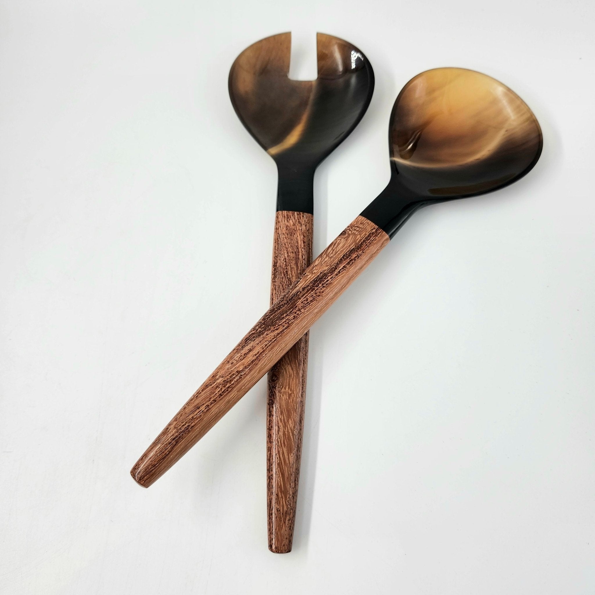 Two wooden serving spoons with glossy tortoiseshell-effect horn heads and medium brown wood handles, crossed on a plain white background.