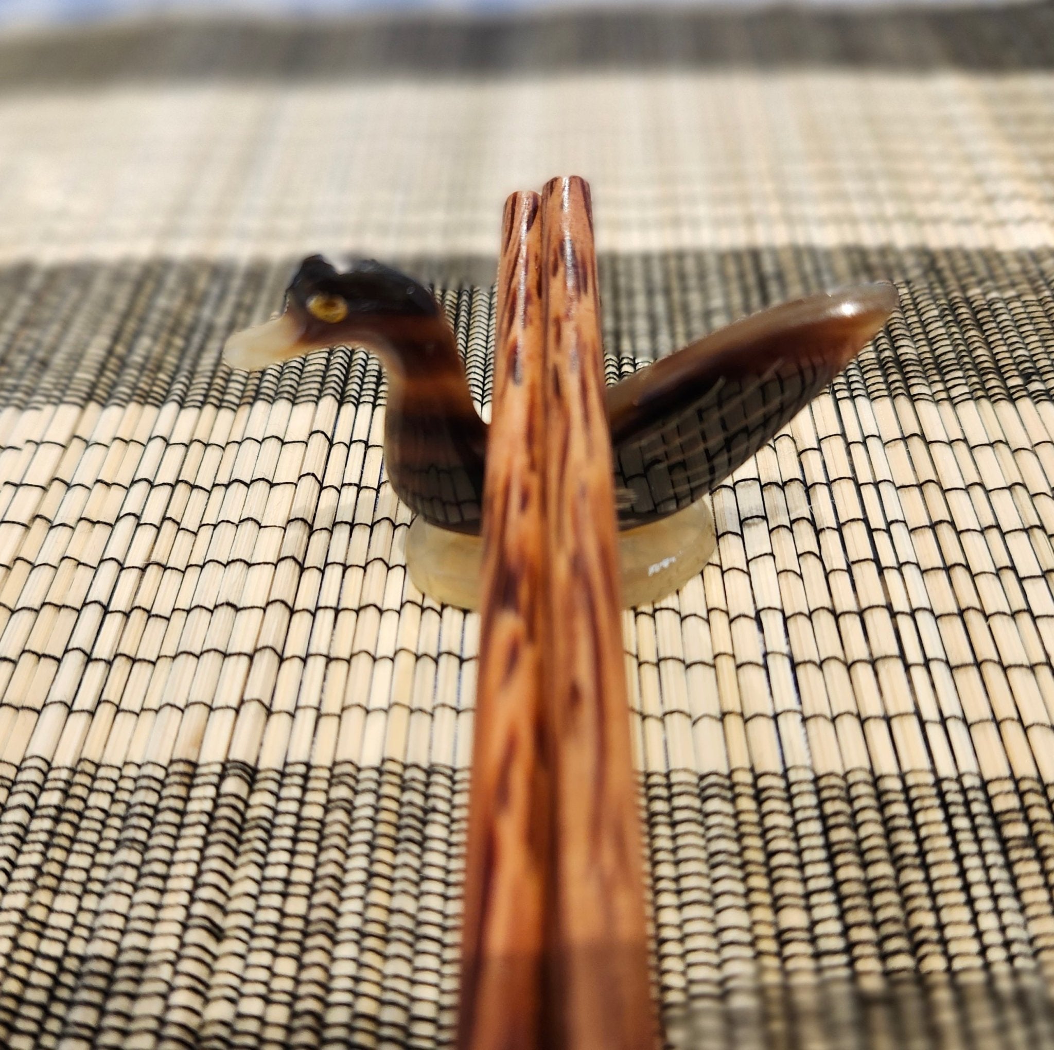A close-up of a small carved duck chopstick stands holding a pair of wooden chopsticks, placed on a woven beige and black mat.