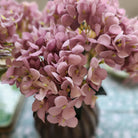 A close-up of a bouquet of dusty pink artificial hydrangea flowers with soft layered petals, arranged in a bronze Salon vase against a softly blurred light green and white background.