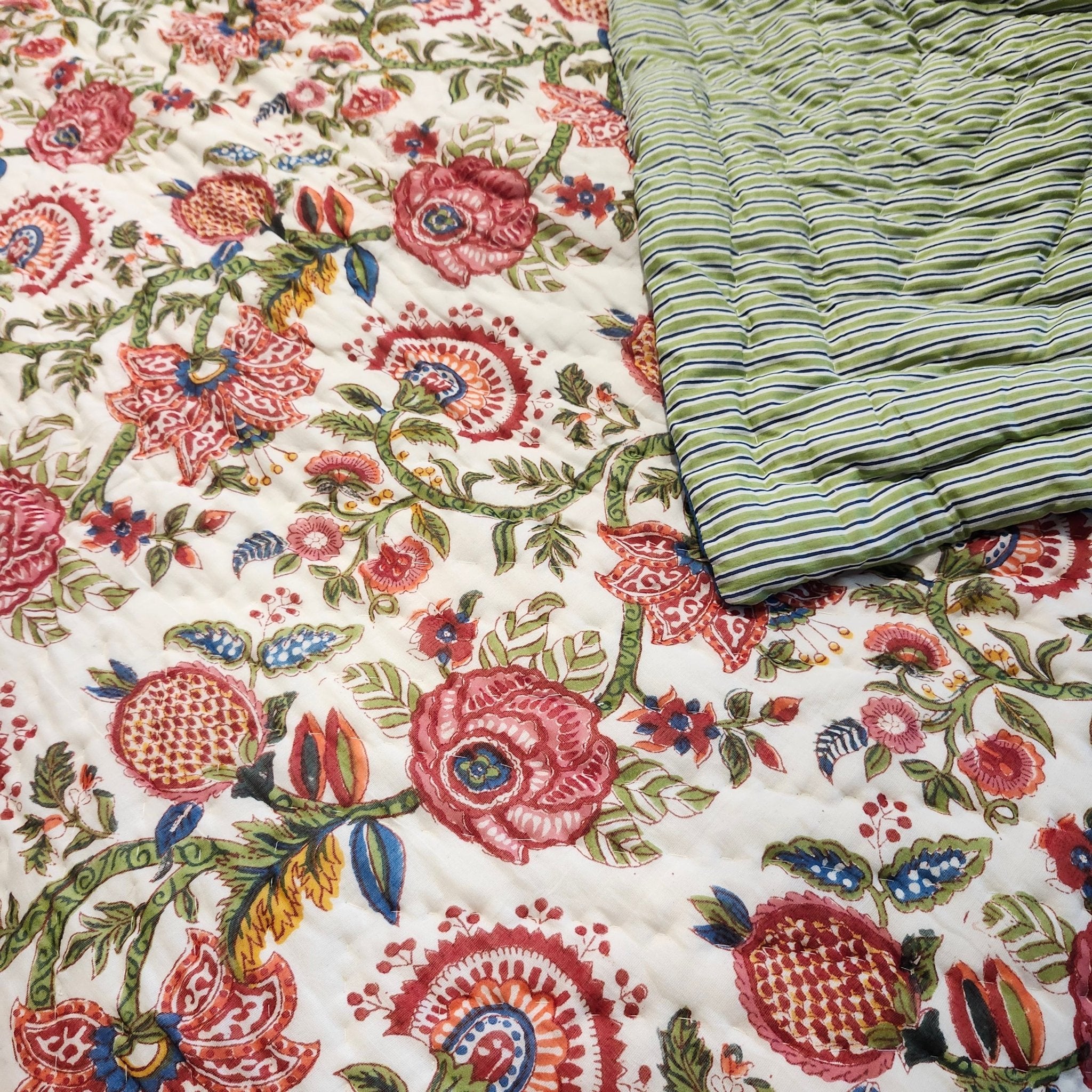A close-up of a quilt with a dense floral and botanical print in pink, red, green, blue, and yellow on an off-white background, with a folded section showing vertical stripes in green, blue, and yellow.