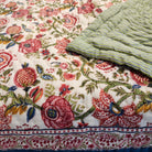 A close-up of a  quilt, showing a detailed floral block-print pattern in pink, red, green, and blue on an off-white background, with a folded section revealing a striped green and blue reverse side.