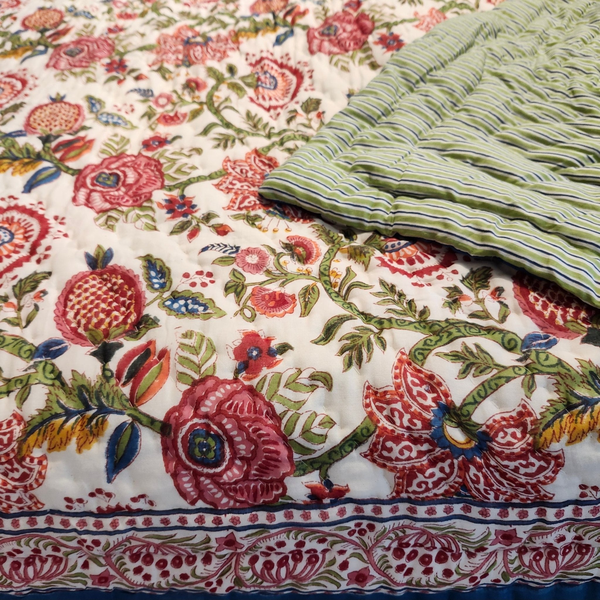 A close-up of a  quilt, showing a detailed floral block-print pattern in pink, red, green, and blue on an off-white background, with a folded section revealing a striped green and blue reverse side.
