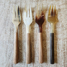 Four decorative forks arranged in a row on a textured light wood surface. The forks have glossy, wooden handles in shades of beige, brown, and dark brown, with smooth, light-colored  horn heads.