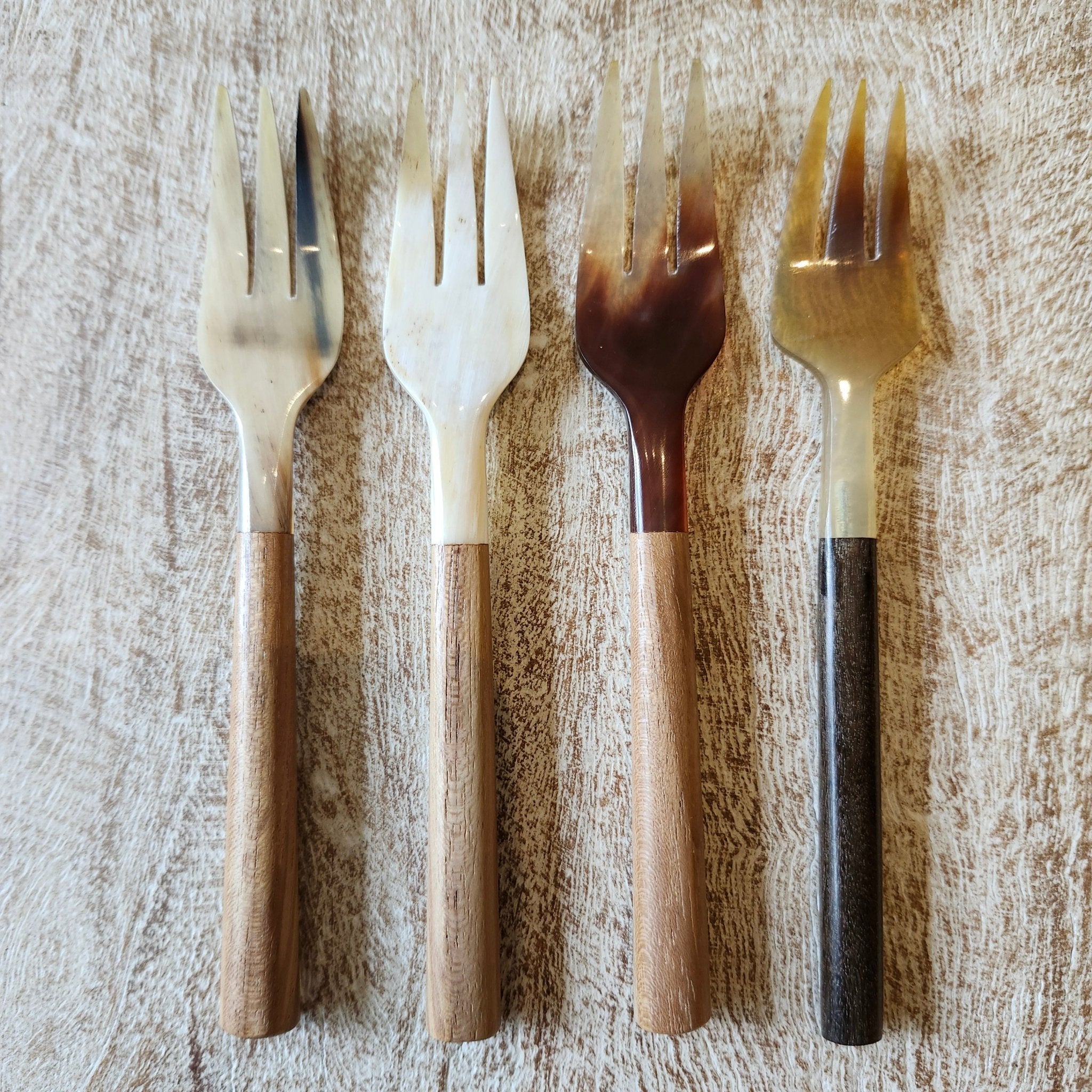 Four decorative forks arranged in a row on a textured light wood surface. The forks have glossy, wooden handles in shades of beige, brown, and dark brown, with smooth, light-colored  horn heads.