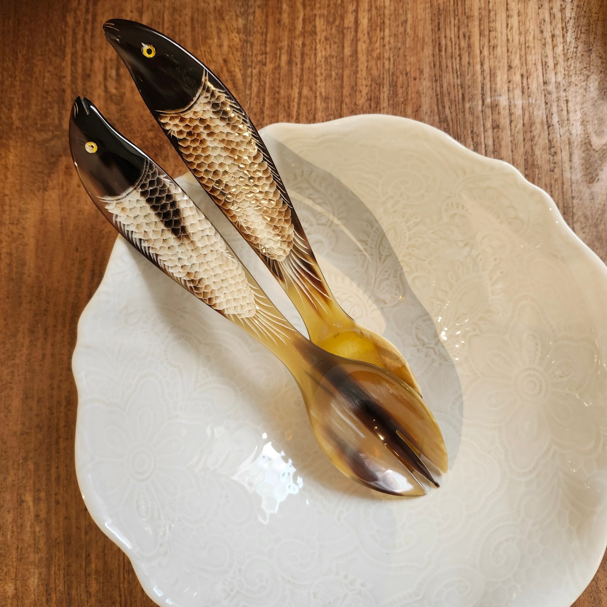 Two decorative fish-shaped salad servers with detailed scales and realistic eyes, resting on a white ceramic plate over a wooden table.